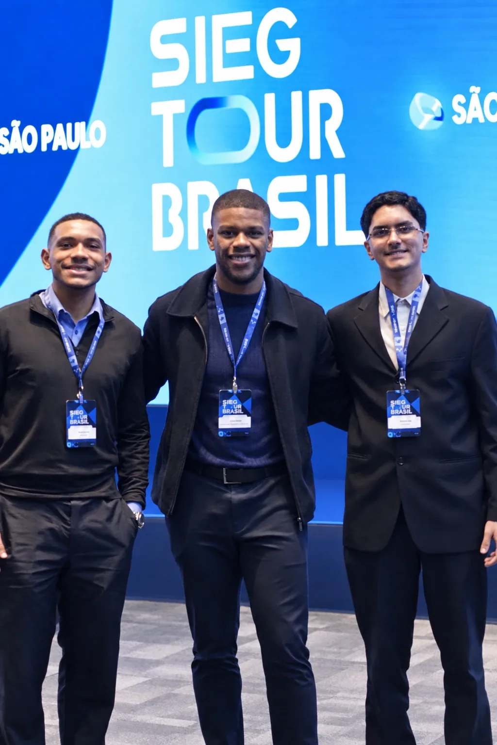 Três participantes com crachá do evento em frente ao painel SIEG Tour Brasil